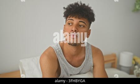 Junger hispanisch-latinoischer Mann auf dem Bett, der seine Wange berührt, während er sitzt und nach unten schaut, in einem Gebäude; nachdenkliche Reflexion. Stockfoto