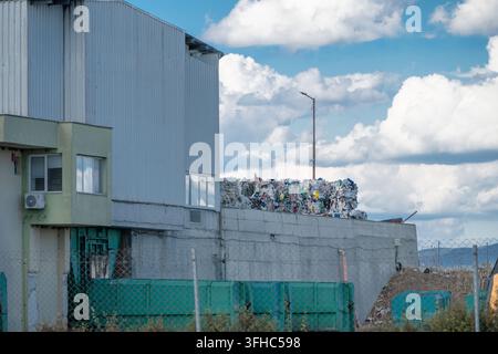 Dichte, gepresste Ballen aus recyceltem Kunststoff und sortiertem Abfall sind auf einer Betonstützmauer neben einer Wellplatten-Industriesortieranlage gestapelt Stockfoto
