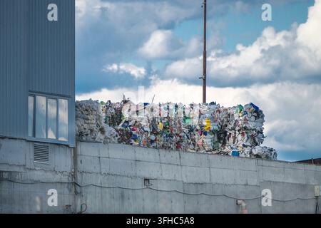 Dichte, gepresste Ballen aus recyceltem Kunststoff und sortiertem Abfall sind auf einer Betonstützmauer neben einer Wellplatten-Industriesortieranlage gestapelt Stockfoto