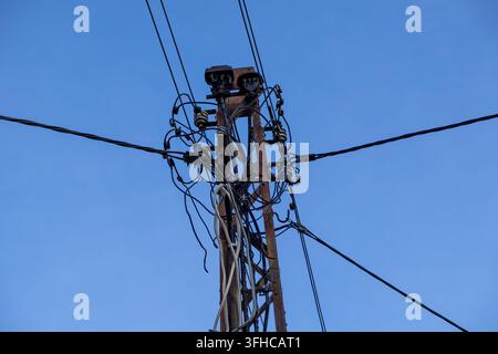 Niedrigwinkelaufnahme eines rostigen Strommastes mit verworrenen Stromleitungen und Keramikisolatoren vor einem klaren blauen Himmel, der lebenswichtige städtische Energieversorgung betont Stockfoto