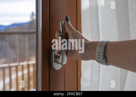 Nahaufnahme einer Frauenhand mit stilvollem grünem Nagellack, die nach einem silbernen Türgriff greift, um eine Glastür zum Balkon in Richtung Bergland zu öffnen Stockfoto