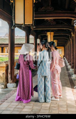 Hue, Vietnam – 9. September 2026. Drei Frauen in farbenfroher traditioneller Kleidung gehen entlang eines historischen Holzgangways, der mit hängenden Laternen gesäumt ist. Stockfoto