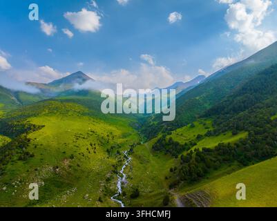 Die Luftperspektive zeigt ein atemberaubendes Bergtal, das in Tageslicht getaucht ist. Ein klarer, flacher Fluss schlängelt sich durch die lebendig grüne Landschaft, flankiert Stockfoto