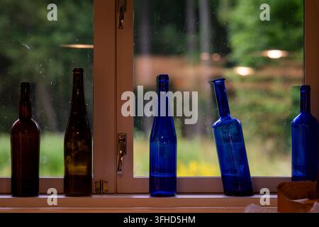 Eine Sammlung leerer Glasflaschen in Braun und Kobaltblau, die auf einer Fensterbank angeordnet sind, mit einem üppigen Garten, der im Hintergrund weich verschwommen ist. Stockfoto