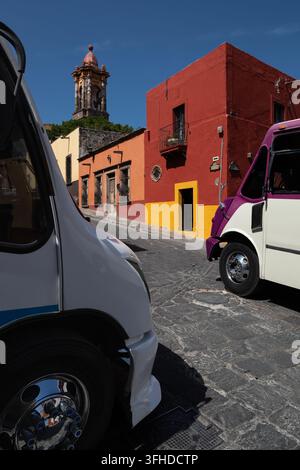 Zwei Busse rahmen eine farbenfrohe koloniale Straße und einen Glockenturm in San Miguel de Allende, Guanajuato, Mexiko ein. Stockfoto