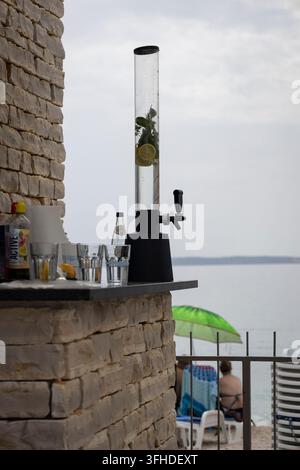 Hoher durchsichtiger Getränkespender gefüllt mit Zitronenscheiben und frischer Minze auf einem Außentresen mit Blick auf das Meer. Stockfoto
