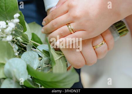 romantisches Paar, das sich an den Händen hält, mit Eheringen in Gold und Weiß vor Grün und Blumen, Nahaufnahme von oben in einer Außeneinrichtung mit Metall Stockfoto