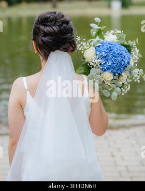 Braut hält einen Blumenstrauß, im Freien in der Nähe eines Gewässers, mit Hochzeitskleid, Schleier, Blumenstrauß, aus einer Perspektive hinter dem Motiv, mit Weiß Stockfoto