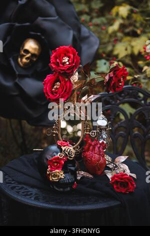 Elegante dunkle Halloween-Komposition mit schwarzen Rosen, Schädeln, roten Blumen und Vintage-Tischgeschirr in einer Waldkulisse. Perfekt für gotische Themen, Party d Stockfoto
