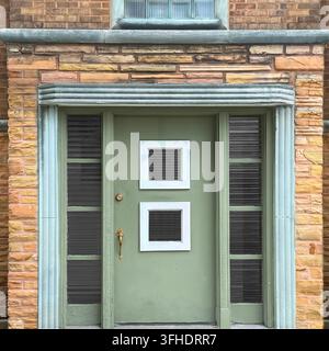 Mid-Century-Tür in Salbeigrün mit zwei quadratischen Fenstern, flankiert von Seitenlichtern in einer abgerundeten Steinverkleidung. Stockfoto