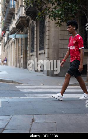 Ein junger Mann in einem roten Sporttrikot überquert eine Straße in der Nähe historischer Architektur in Barcelona, Spanien. Stockfoto