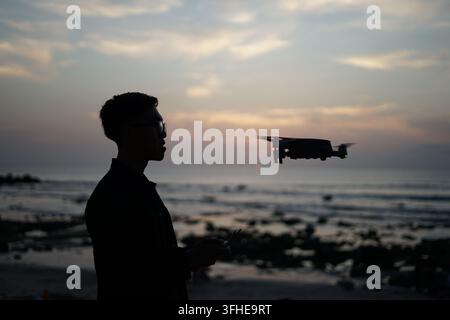 Seitenansicht-Silhouette eines jungen Mannes, der eine Drohnenkamera am Strand bei Sonnenuntergang bedient Stockfoto