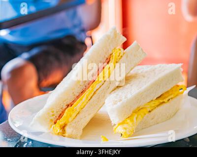 Zwei dreieckige Schinken-Eier-Sandwiches ruhen auf einem weißen Teller, während Gäste im sanft verschwommenen Café-Hintergrund entspannen. Stockfoto