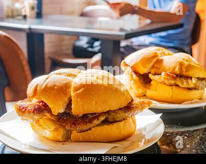 Knusprige Schweinekoteletts, die zwischen weichen Brötchen liegen, stehen auf weißen Tellern an einem geschäftigen Esstisch mit einem Kunden im Hintergrund. Stockfoto