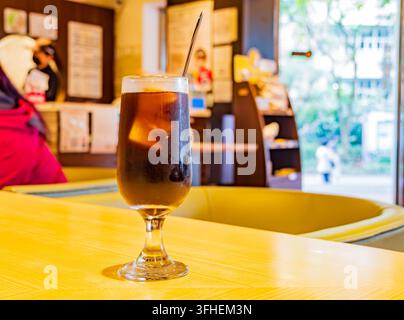 Taipei, Taiwan, 14. APR 2024 – Ein hohes Glas dunkler Eistee mit einem Metallstrohhalm steht auf einer hellgelben Holzoberfläche in einem Café. Stockfoto