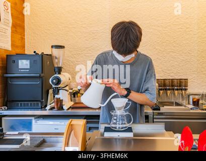 Macau, China, 15. APR 2024 – Ein konzentrierter Barista, der eine Maske trägt, gießt heißes Wasser aus einem weißen Schwanenhalskessel auf Kaffeemehl in einem Glas-Dripper. Stockfoto