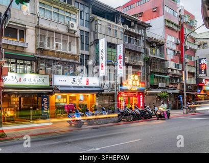 Taipei, Taiwan, 19. APR 2024 – Eine belebte Straße in Taipei bei Nacht zeigt leuchtende Restaurantschilder, aufgereihtes Motorradverkehr und Langzeitbelichtungs-Lichtspuren auf Stockfoto