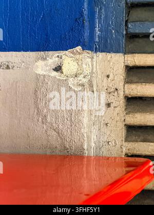 Nahaufnahme einer gealterten blau und cremefarben gestrichenen Wand mit rauem Putz, Lüftungsschlitzen und einem hellroten Vordergrund-element. Stockfoto