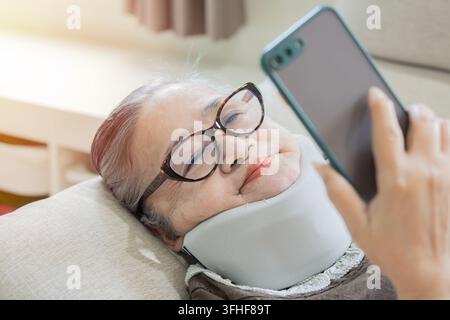 Eine ältere asiatische Frau, die einen Halsstützkorsett trägt, nimmt ein Selfie oder führt einen Videoanruf auf ihrem Smartphone, während sie bequem liegt. Stockfoto