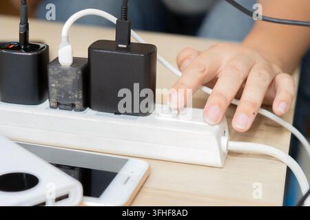 Nahaufnahme einer Hand, die einen weißen Verlängerungskabelschalter bedient, der auf einem Schreibtisch mit mehreren schwarzen Ladegeräten und USB-Kabeln überfüllt ist. Stockfoto