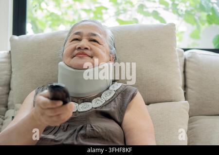 Eine ältere asiatische Frau, die einen Zervikal-Kragen trägt, sitzt bequem auf einer Couch und zeigt mit einer Fernbedienung auf den Fernseher, während sie zu Hause entspannt Stockfoto
