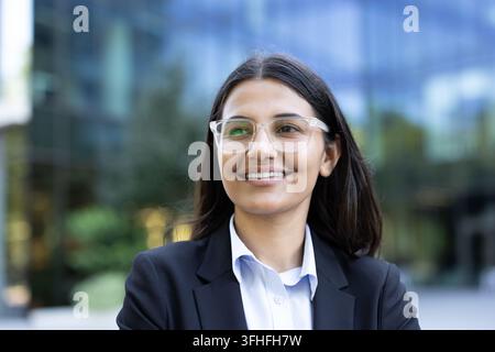 Professionelle Frau im Business-Anzug lächelt, Konzept für Unternehmenserfolg und Führung, junge Unternehmerin mit Brille steht in einem modernen Büroviertel. Selbstbewusstes Geschäft. Stockfoto