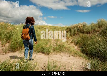Junge Frau mit lockigem Haar trägt eine lässige blaue Jacke und Jeans und geht auf einer Küstensanddüne durch Strandhafer an einem windigen Sonnentag weg. Stockfoto