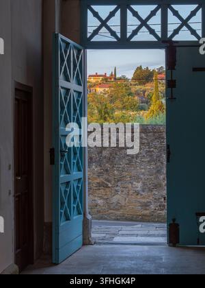 Offene blaue Holztüren mit geometrischen Paneelen rahmen eine Steinmauer im Innenhof und einen sonnenbeschienenen Blick auf das Dorf in der Landschaft im warmen Abendlicht. Stockfoto