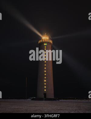 Blick auf den beleuchteten Grotta-Leuchtturm, der einen hellen Strahl in den dunklen Nachthimmel über der verschneiten Landschaft in Island wirft. Stockfoto