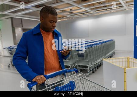 Ein junger schwarzer Mann in einer blauen Jacke und einem orangefarbenen Hemd überprüft sein Telefon, während er einen Einkaufswagen in einem geräumigen Laden Gang voller Wagen schiebt Stockfoto