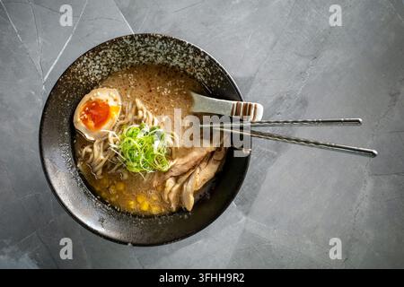 Köstliche Schüssel Ramen-Nudelsuppe serviert mit Hühchenscheiben, Ramen-Ei, Frühlingszwiebeln und Essstäbchen auf einer dunkelgrauen Steinoberfläche. Stockfoto