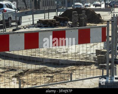 Metallsicherungszäune markieren eine aktive Straßenerneuerungsbaustelle, gefüllt mit losem Sand und gestapelten Pflastersteinen, und heben kommunale Reparaturprojekte hervor Stockfoto