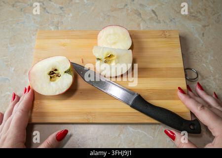 Frische rote Apfelscheiben und ein dunkles Schälmesser liegen auf einem Holzschneidebrett, während die Hände einer Person an den Seiten ruhen, gesehen aus einer Oberblickperspektive. Die Kom  Stockfoto