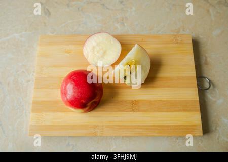 Eine ganze rote Apfelhälfte und frisch geschnittene Viertel liegen auf einem rechteckigen Holzschneidebrett, das auf einer Marmorküchenoberfläche in einer Hochwinkelansicht platziert ist. Die simp Stockfoto