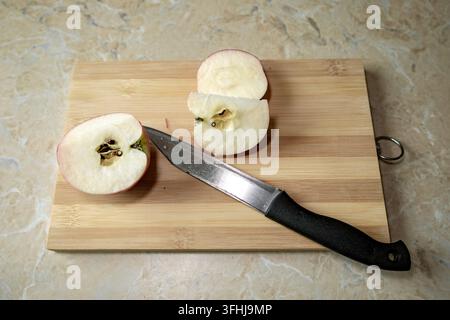Frisch geschnittene Rotapfelstücke liegen neben einem schwarzen Küchenmesser auf einem rechteckigen Holzbrett, das auf einer Marmorarbeitsplatte platziert ist. Die Draufsicht Stockfoto