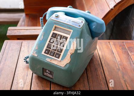 Ein verwitterter türkisfarbener Münzfernsprecher mit abblätternder Farbe steht auf einem Holztisch, sein Hörer ruht auf der Gabel neben einer abgenutzten Zifferntastatur und thailändischem Text Stockfoto