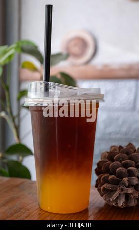 Ein Eiskaffee mit Farbverlauf von Dunkelbraun zu Goldorange steht neben einer Kieferngespinne auf einem Holztisch mit grünen Pflanzen im Hintergrund Stockfoto