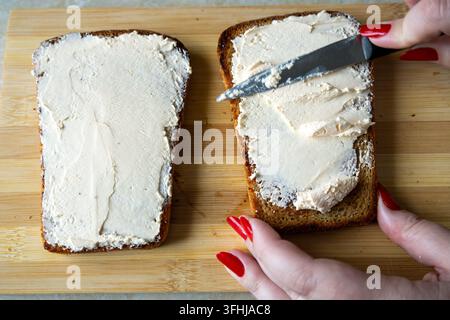 Eine Person streicht weißen Frischkäse mit einem kleinen Messer auf getoastetes Brot auf einem Holzschneidebrett. Hände halten die Scheibe während der Zubereitung. Stockfoto