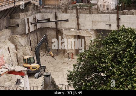 Tiefer Aushub zeigt Stützmauern und vertikale Stahlbügel in einem hohen Graben, mit Holzschalung, Gerüst und Bagger, der unter Staub arbeitet Stockfoto