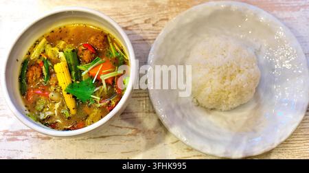 Eine köstliche Schüssel japanischer Suppencurry gefüllt mit bunten Gemüsen, Mais, Fleischbällchen und einer Beilage aus weißem Reis, serviert auf einem rustikalen Holztisch. Stockfoto