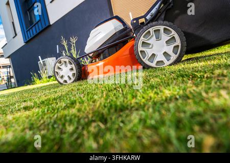 Ein Rasenmäher mit Grash Auffangsack steht auf einem Rasen neben einem Haus. Die Nahaufnahme zeigt seine Räder und den Schneidbalken. Stockfoto
