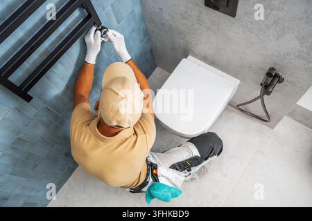 Luftbild eines Arbeiters, der die elektronische Heizungsregelung an einem Handtuchheizkörper neben einer wandmontierten Toilette in einem gefliesten Badezimmer einstellt. Stockfoto