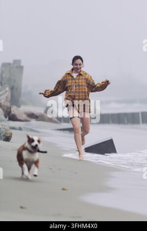Junge erwachsene Frau mit dunklen Haaren geht barfuß neben einem verspielten Hund entlang eines nebligen Strandes, trägt ein kariertes Hemd und Shorts in der Nähe von Wellen, Felsen und Küstenkuchen Stockfoto