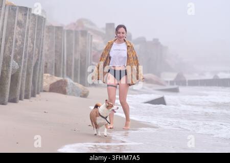 Junge erwachsene Frau mit dunklen Haaren geht barfuß neben einem kleinen braun-weißen Hund entlang eines nebligen Strandes, trägt ein offenes kariertes Hemd in der Nähe von blassem Schaum und Beton Stockfoto