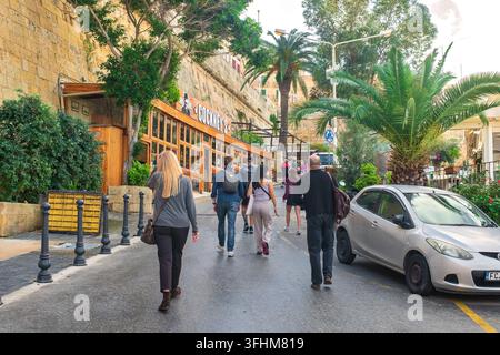 Valletta, Malta - 2. Dezember 2025: Menschen gehen in der Stadt entlang einer malerischen Straße an einem Restaurant, einer historischen Steinbefestigungsmauer, Palmen vorbei Stockfoto