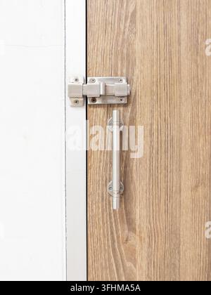 Nahaufnahme eines modernen Edelstahl-Schiebebolzenverschlusses und Griffs, montiert auf einer Holztür mit Maserung neben einer weißen Wand. Stockfoto