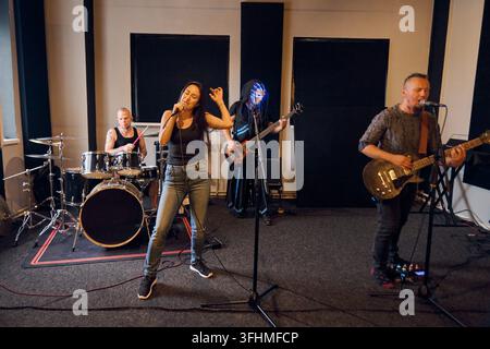 Junge erwachsene Frau mit dunklen Haaren singt in ein Mikrofon neben einem männlichen Schlagzeuger, einer weiblichen Bassistin und einem männlichen Gitarristen in einem schallgedämmten Studio mit warmem Innenlicht Stockfoto