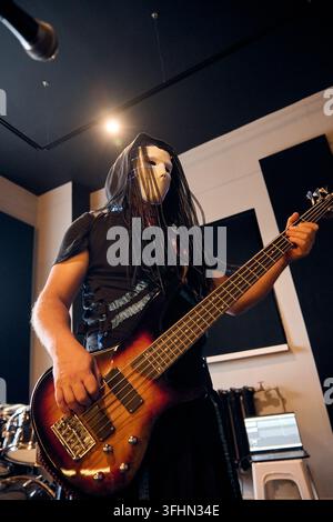 Junge Frau mit langen dunklen Haaren spielt Sunburst-Bassgitarre im Aufnahmestudio, unter warmer Deckenbeleuchtung und neben Akustikpanelen, Trommeln und Audio Stockfoto