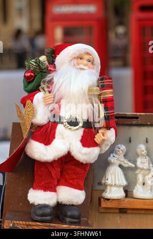 Weihnachtsmann-Spielzeugfigur im traditionellen roten Anzug, die eine Geschenkschachtel und eine Tasche auf einem hölzernen Marktstandregal hält, mit unscharfem Hintergrund aus roten Telefonzellen. Stockfoto