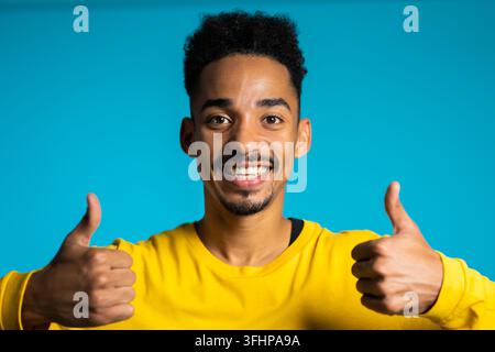 Attraktiver schwarzer Mann in gelber Kleidung auf blauem Studiohintergrund lächelt in die Kamera und gibt Daumen hoch. Glücklicher Typ zeigt Geste eines Stockfoto